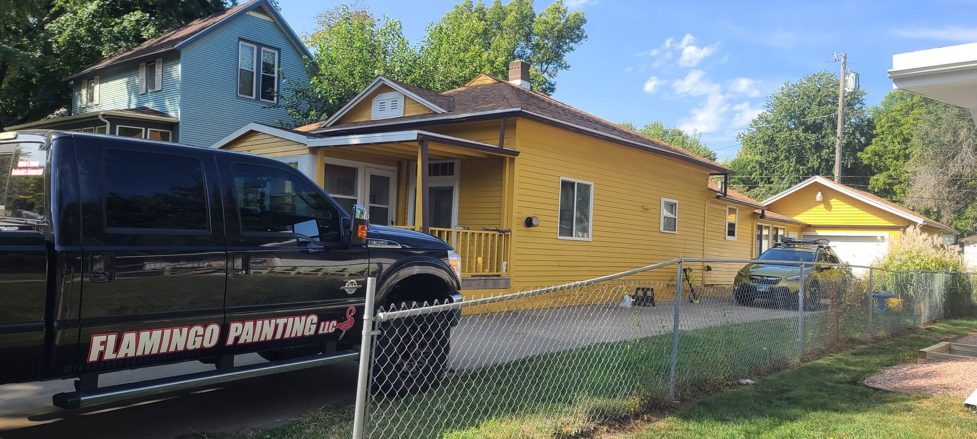 A yellow house with a black pickup truck parked in front of it. Blue house is visible in the background.