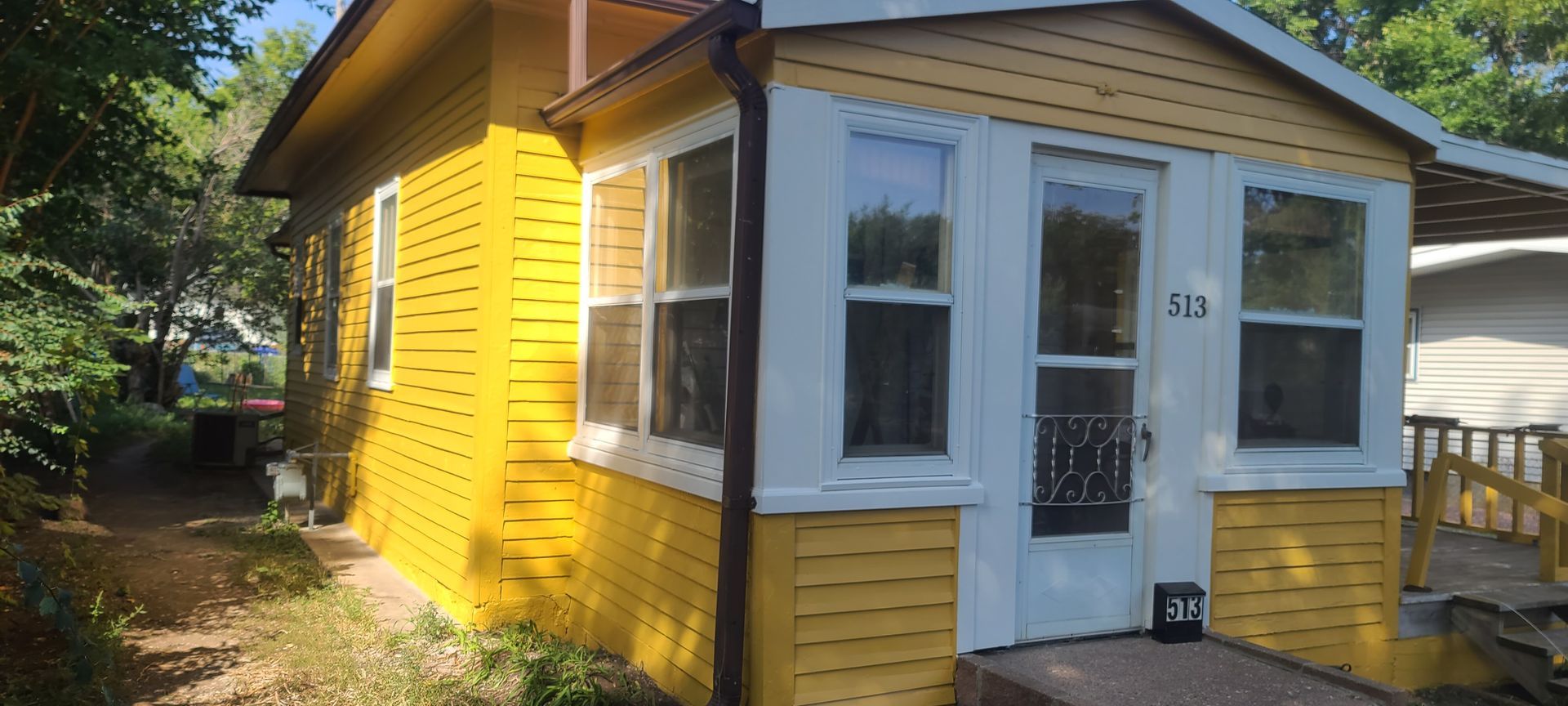 Yellow house with white trim, windows and a door. There is a path and greenery in front.