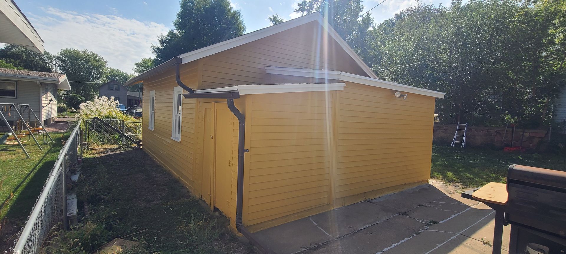 Yellow building with white trim, brown gutters, and a gray patio. Trees and green grass are in the background.