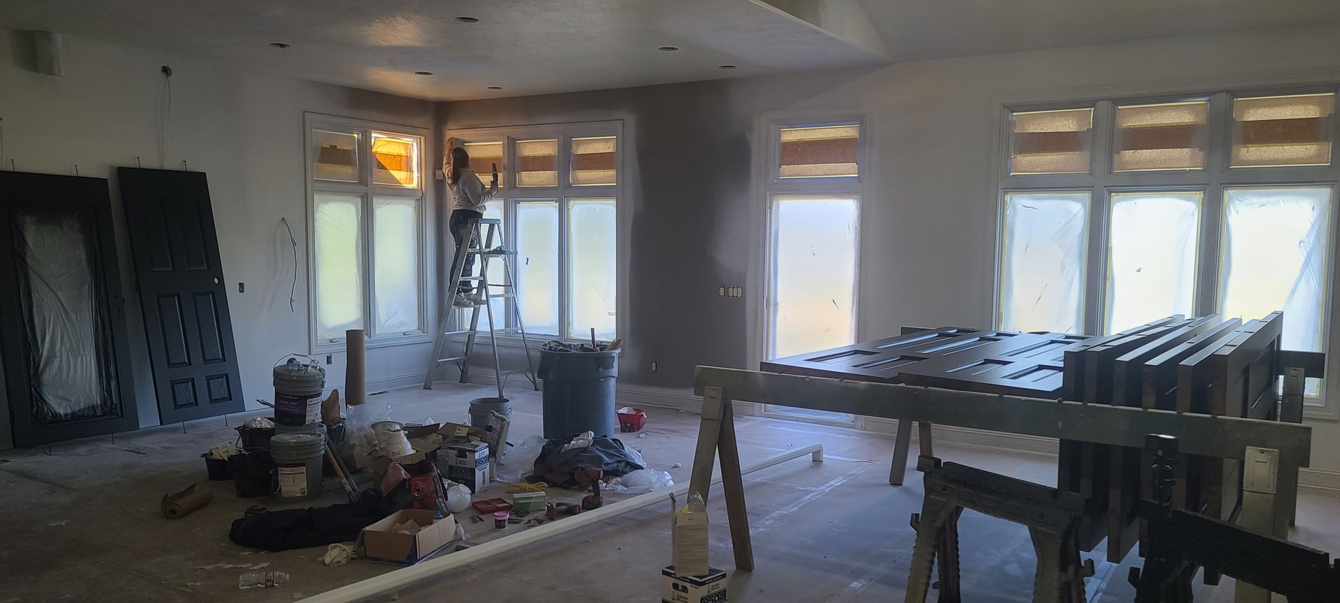 Interior of a room under construction with a person on a ladder working on the windows.