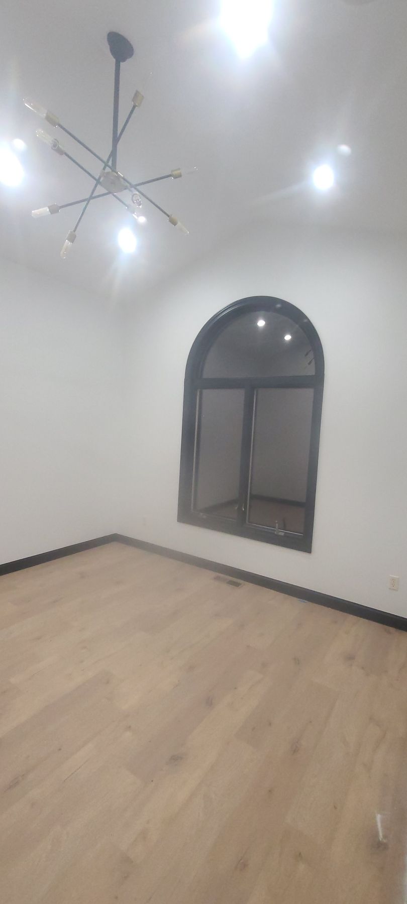 Empty room with wooden floors, arched window, and modern chandelier. Walls are white.