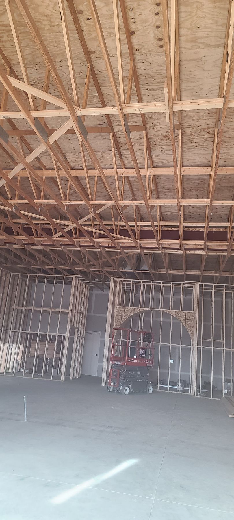Interior view of a building under construction. Wooden framing and roof structure are visible. There is a red machine at the center.