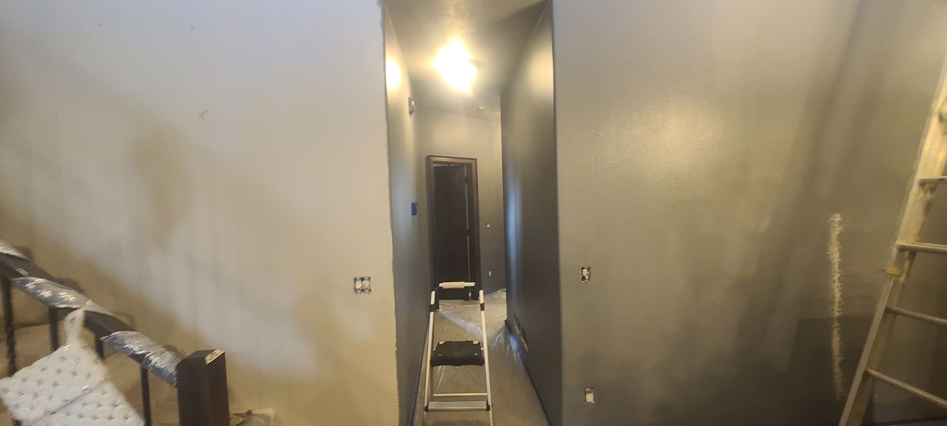A narrow hallway with gray walls, a closed dark door, and a stepladder. Staircase on the left, wooden ladder on right.