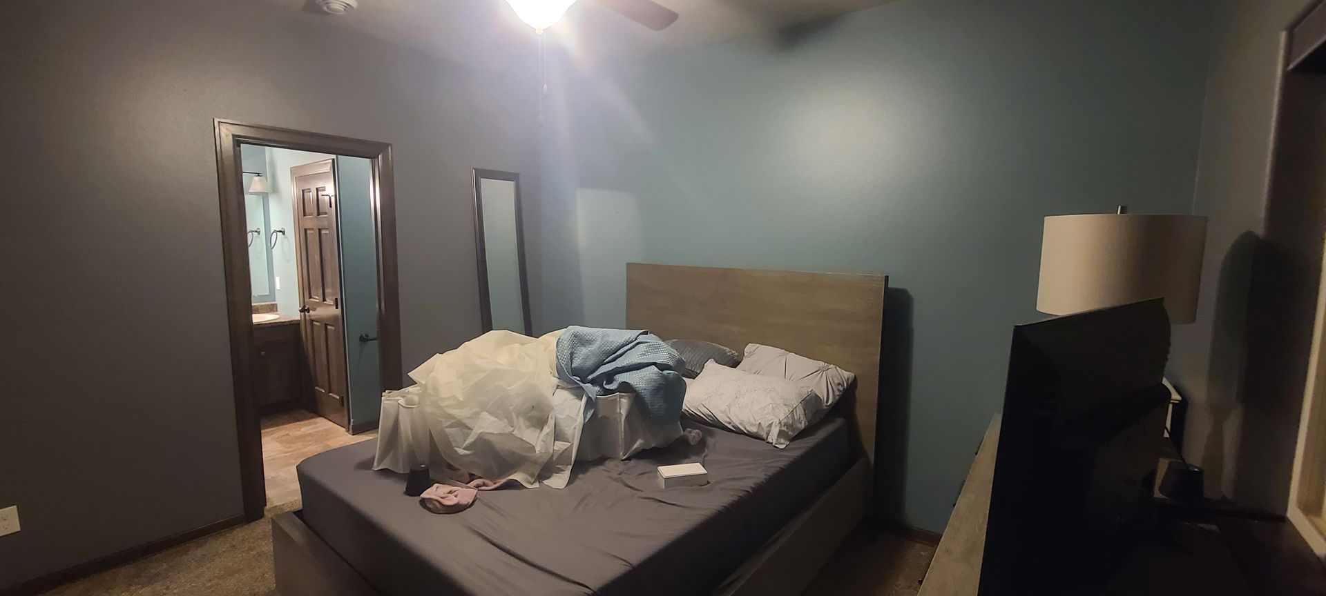 A messy bedroom with a bed, dresser, and an open doorway to a bathroom. The walls are blue and gray.