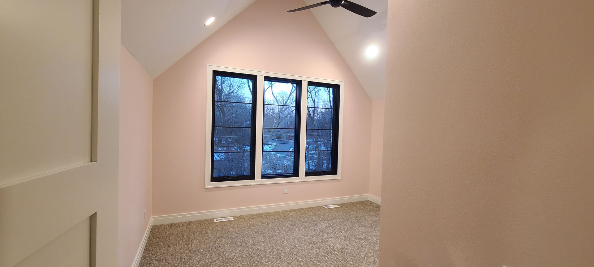 A room with three windows and a sloped ceiling. The walls are pink and the floors are carpeted.