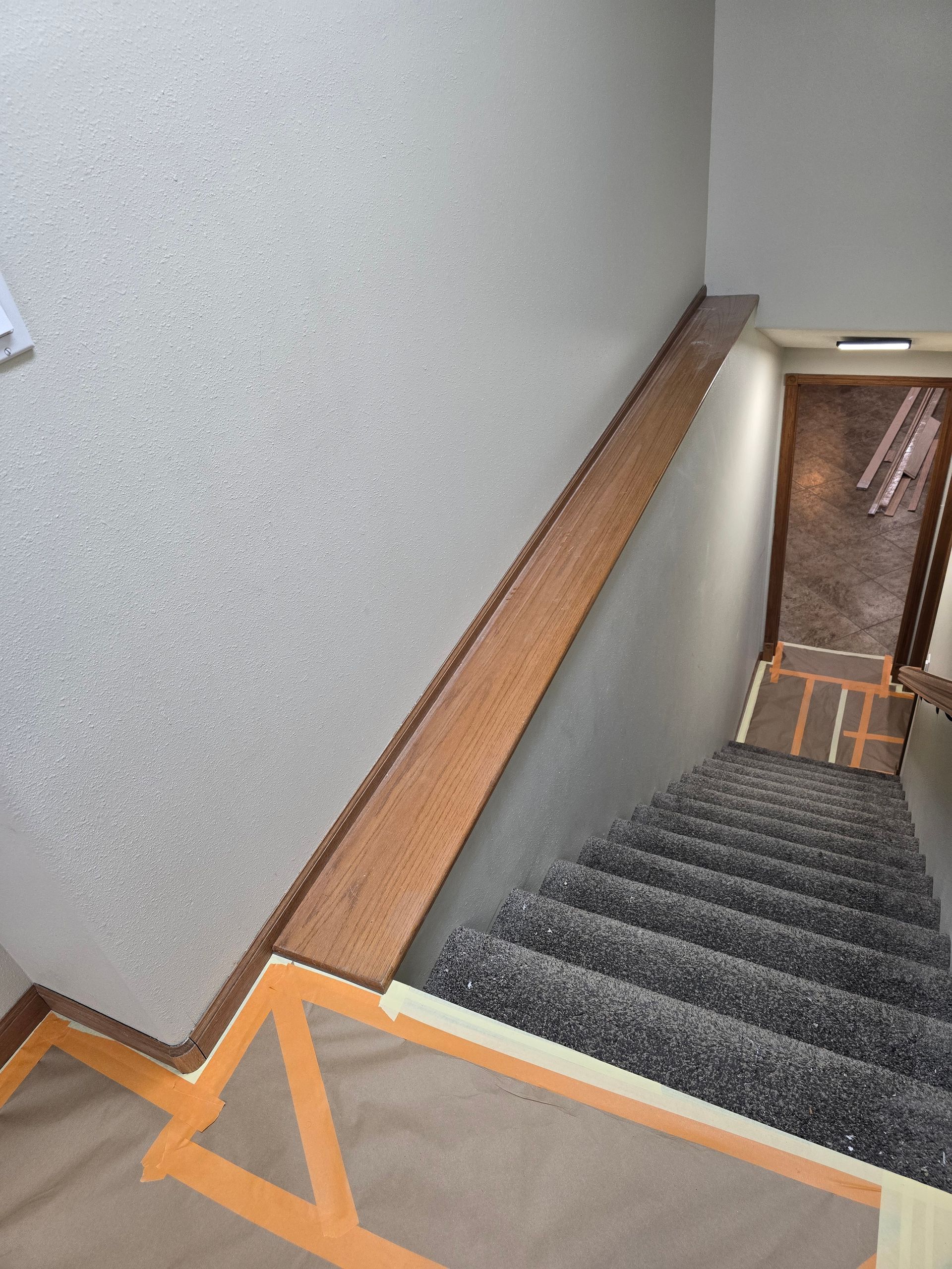 Staircase with gray carpet, brown wooden handrail, and off-white walls. Orange tape protects carpet edges.