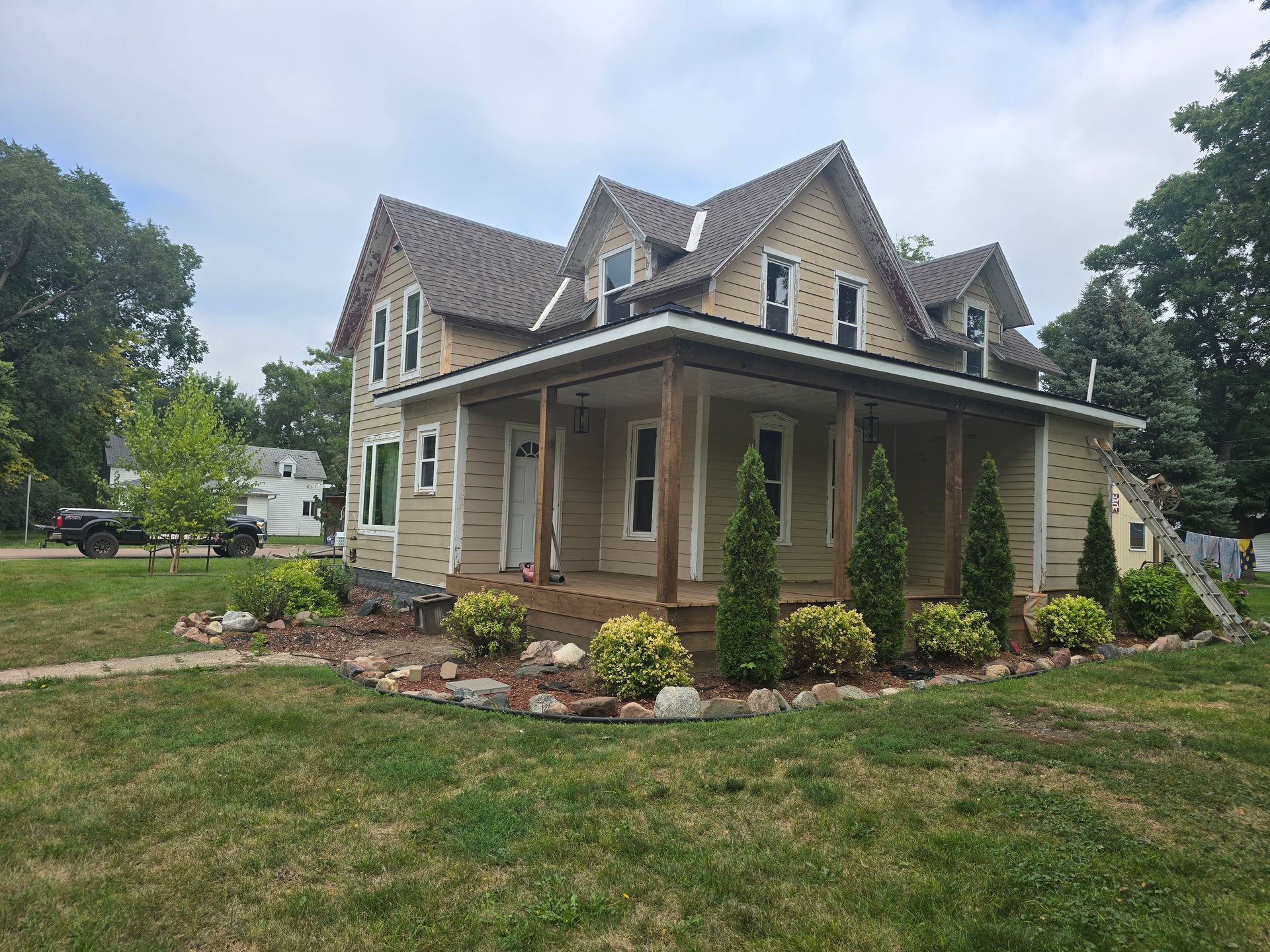 Two-story house with a wraparound porch, tan siding, and brown roof. Landscaped front yard with shrubs and a rock border.