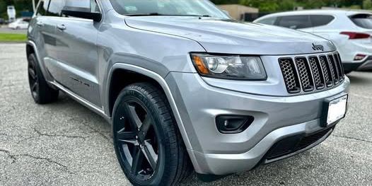 Silver Jeep Grand Cherokee with black wheels parked outdoors on a gray surface.