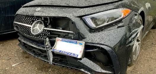 Black Mercedes with front-end damage; damaged grill, license plate, and bumper, appears to be on wet pavement.