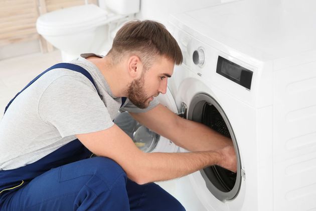 Technician in blue overalls repairing a front-loading washing machine in a white laundry room