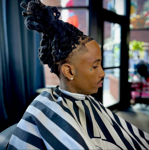 Man with locs and a tapered haircut, wearing a barber's cape, indoors.