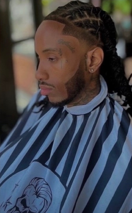 Man with braided hair and a patterned barber cape, looking down.