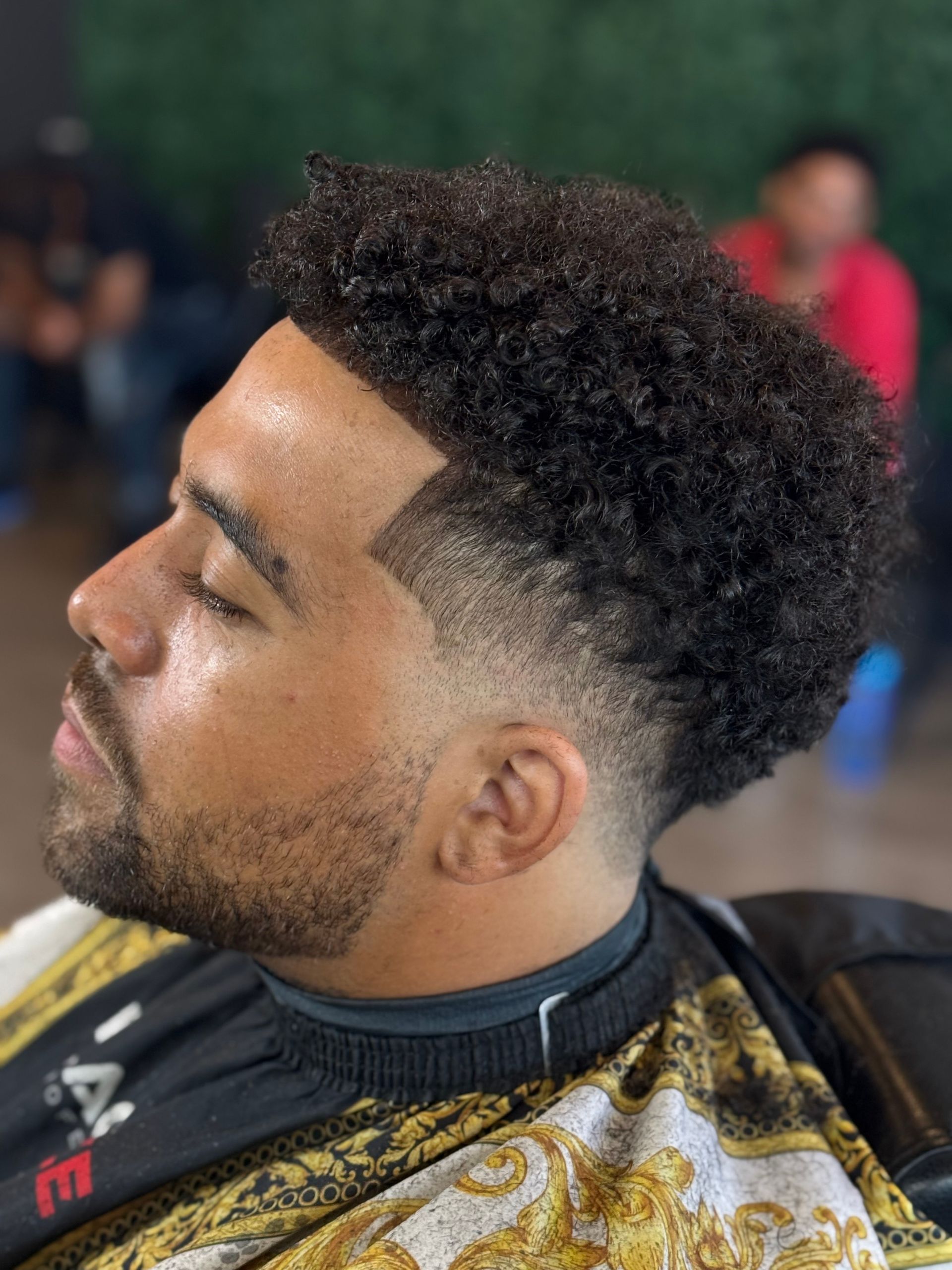 Man with dark curly hair and a skin fade haircut; seated in a barber shop, looking to the side.