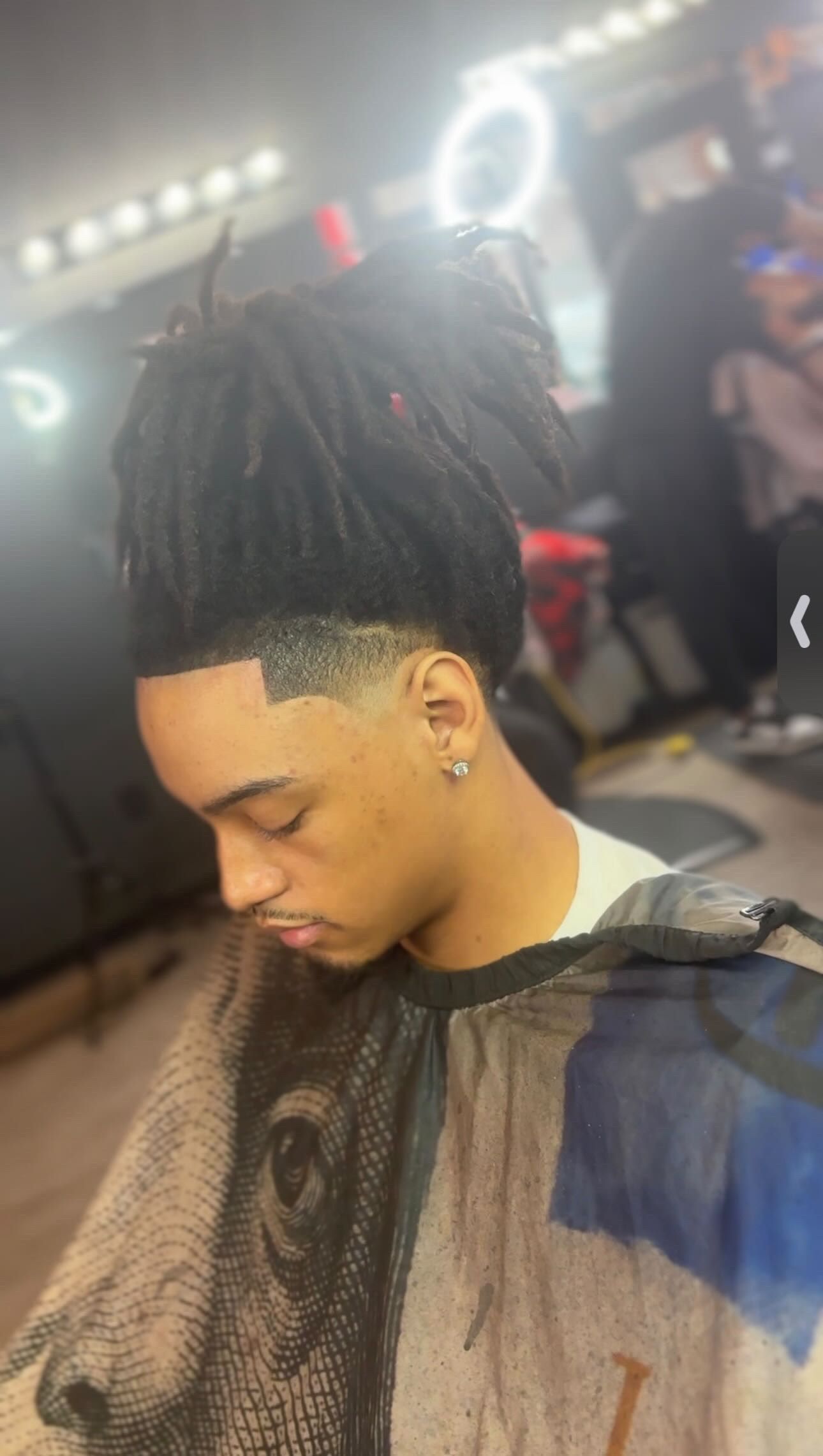 Person with a faded haircut and dreadlocks, wearing a barber cape, in a barbershop.