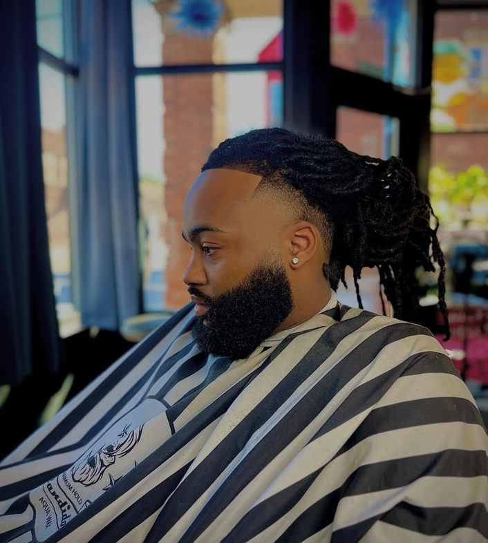 Man with beard and dreadlocks in a barber cape, hair styled, side profile in a salon.