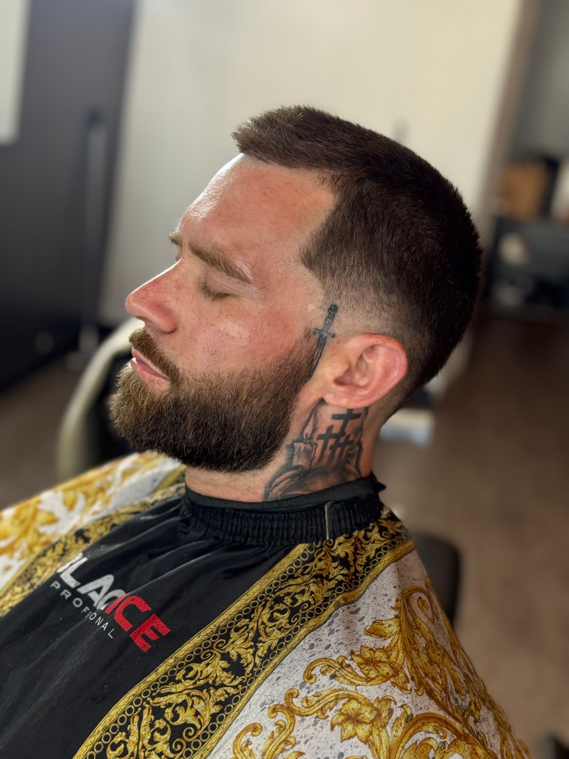 Man with dark hair and a beard, in a barber's chair; fresh haircut and neck tattoo visible.