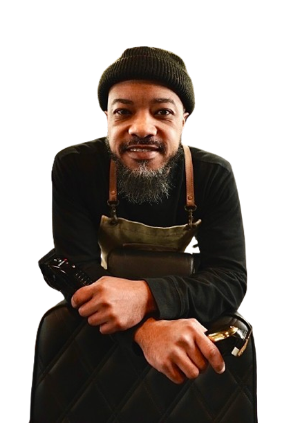 A man with a beard wearing a hat and apron is sitting on a suitcase.