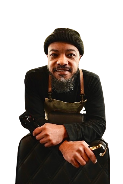 A man with a beard wearing a hat and apron is sitting on a suitcase.