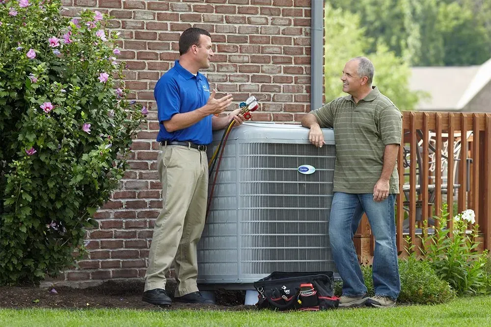 HVAC technician shows a homeowner an air conditioner unit outside a brick house.