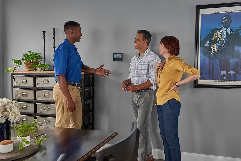 HVAC technician talks to a couple near a thermostat on a wall. Indoor setting, they appear to be discussing.