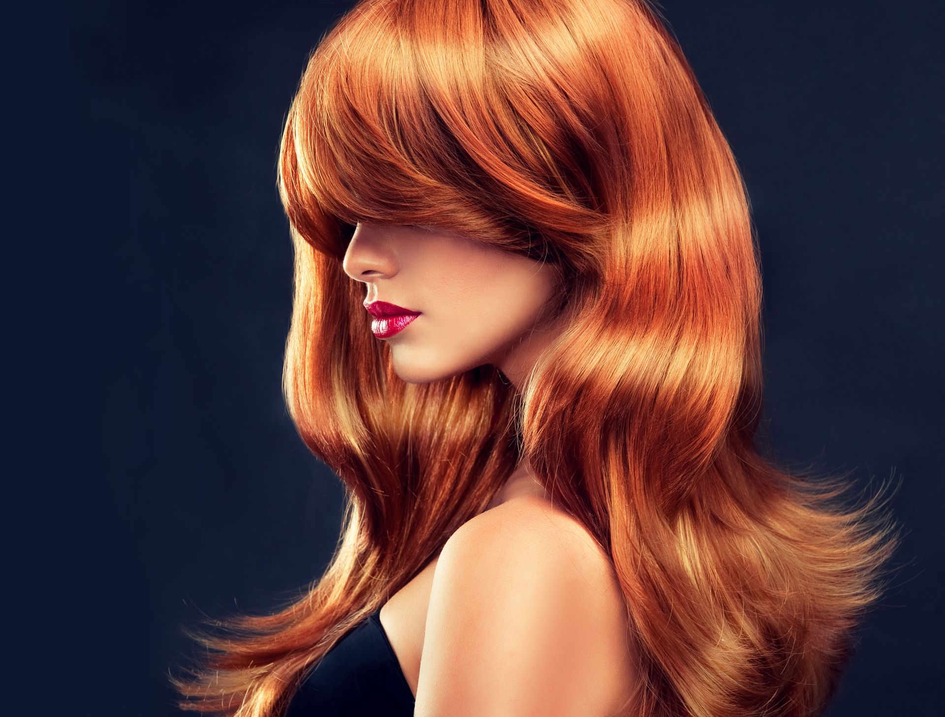 A woman with long red hair and red lips is wearing a black top.