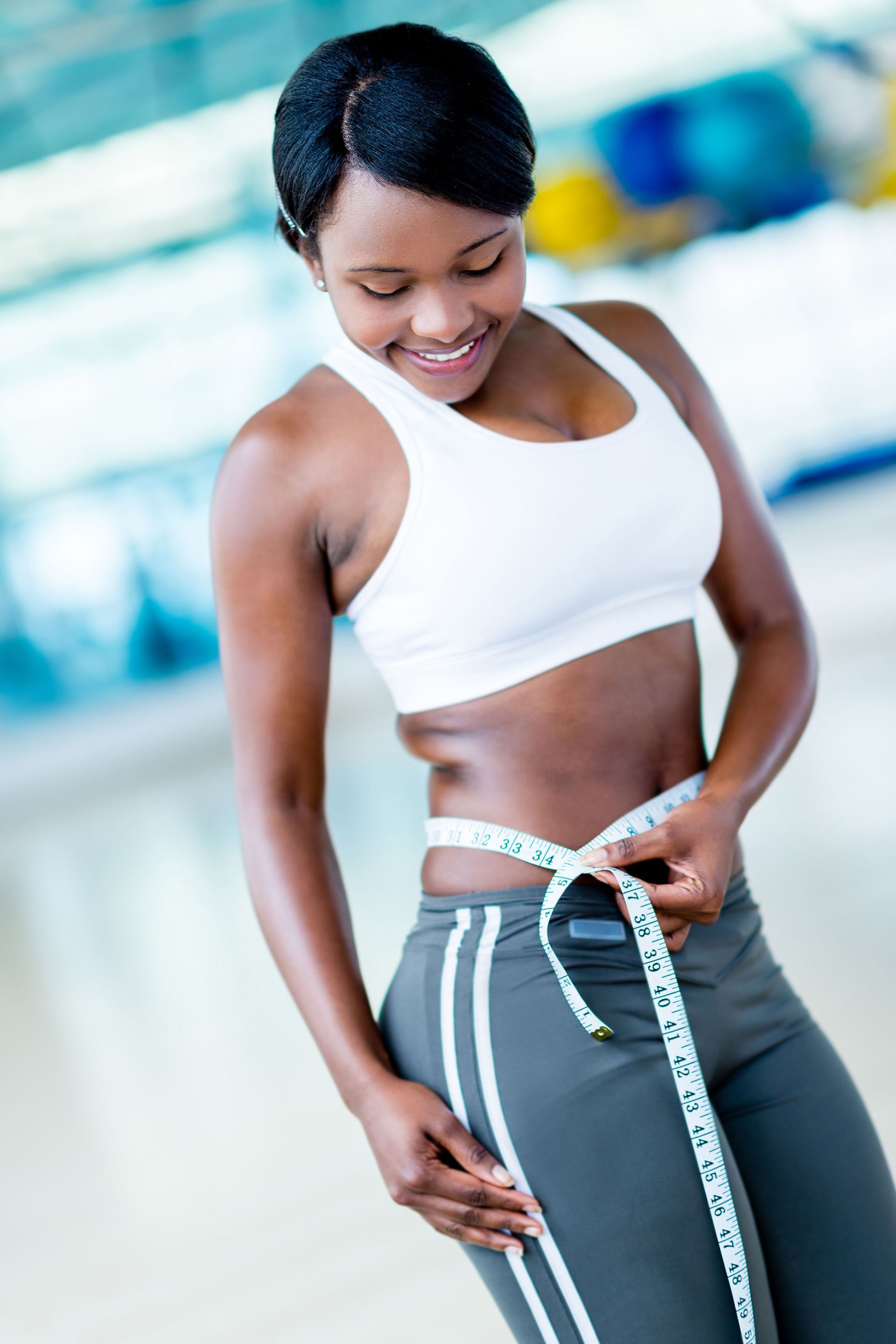 Woman in workout clothes measures her waist with a tape measure, smiling.