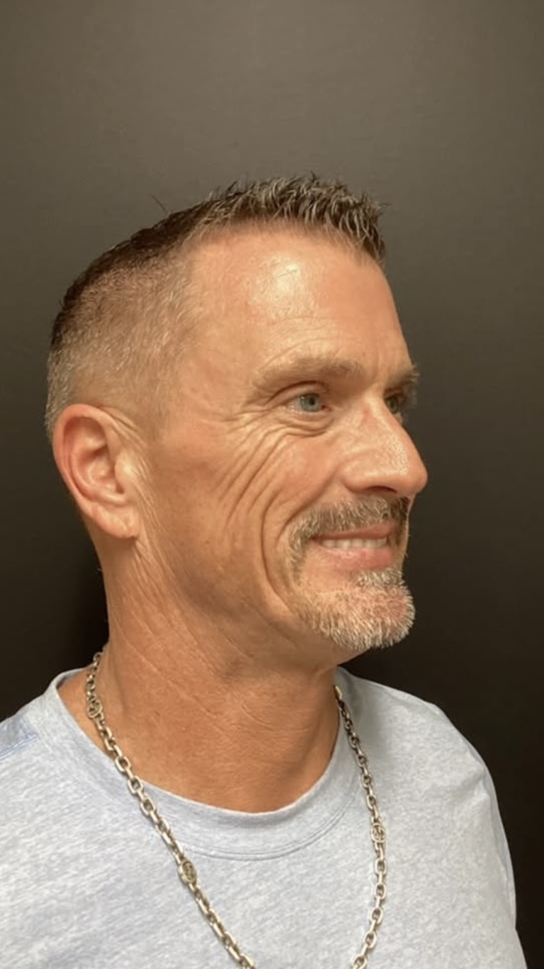 Man with short, textured hair and goatee, smiling, wearing a light blue shirt and necklace, against a dark background.