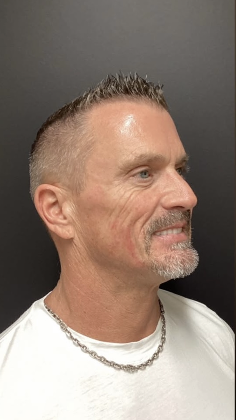 Man with short, spiky hair and a trimmed goatee smiles. Wearing a white shirt and silver necklace.