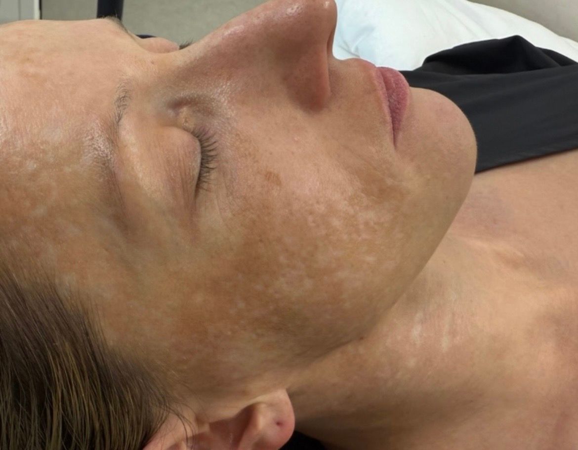 Woman's face close-up, eyes closed, with a light-colored cream applied, lying on a white surface. 