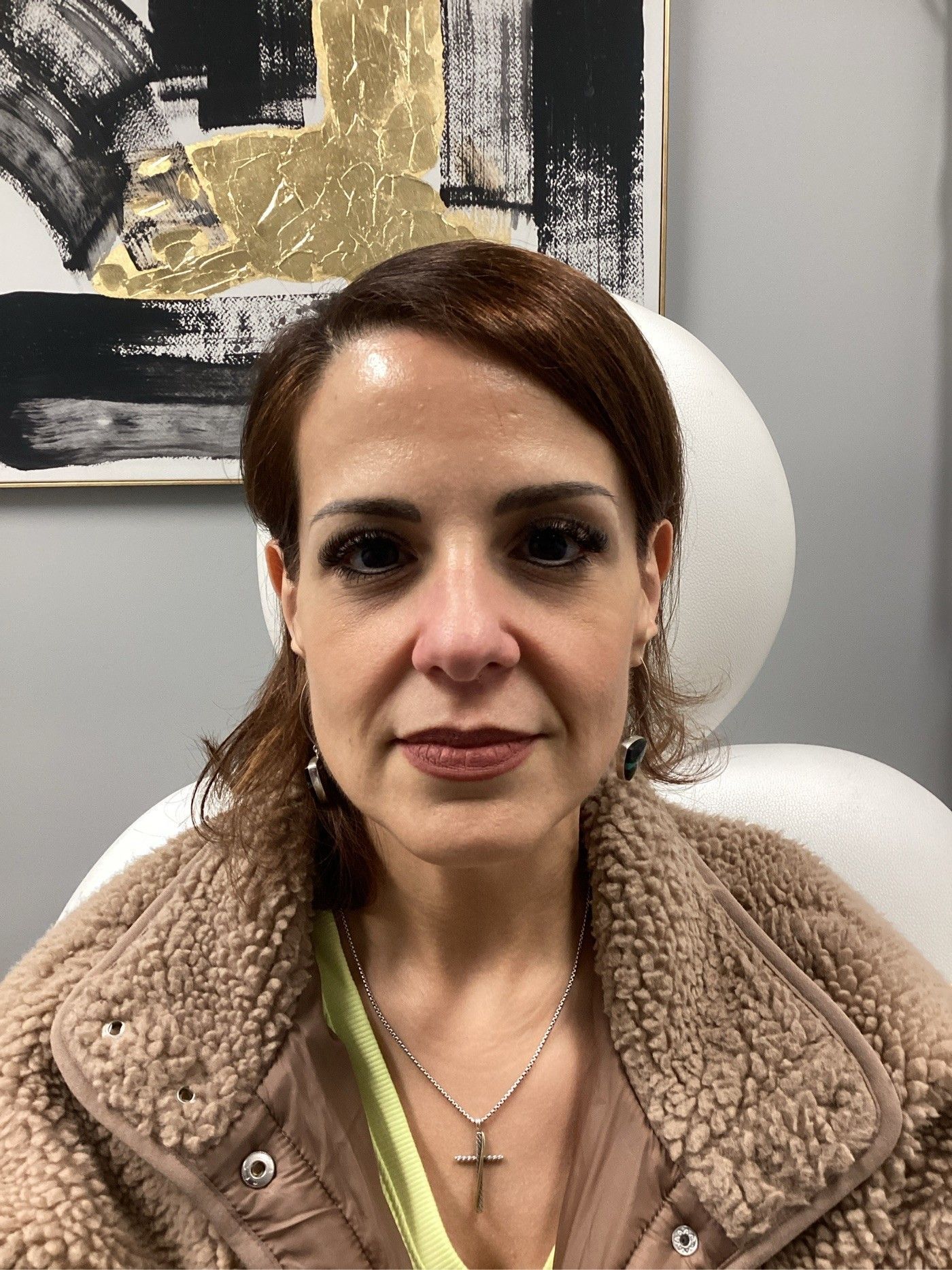 Woman with brown hair, wearing a brown jacket, sitting in a chair.  A painting hangs on the wall behind her.