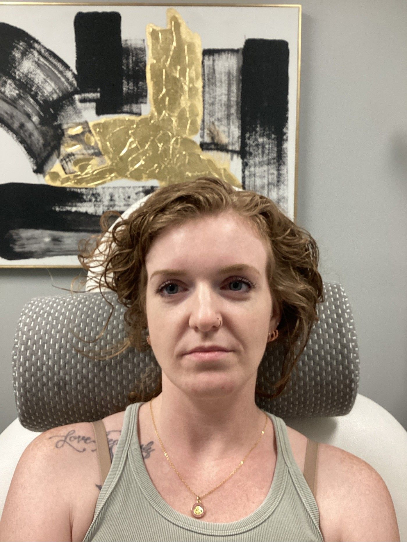 Woman with curly auburn hair in a tank top, sitting against a grey chair. Abstract painting in the background.