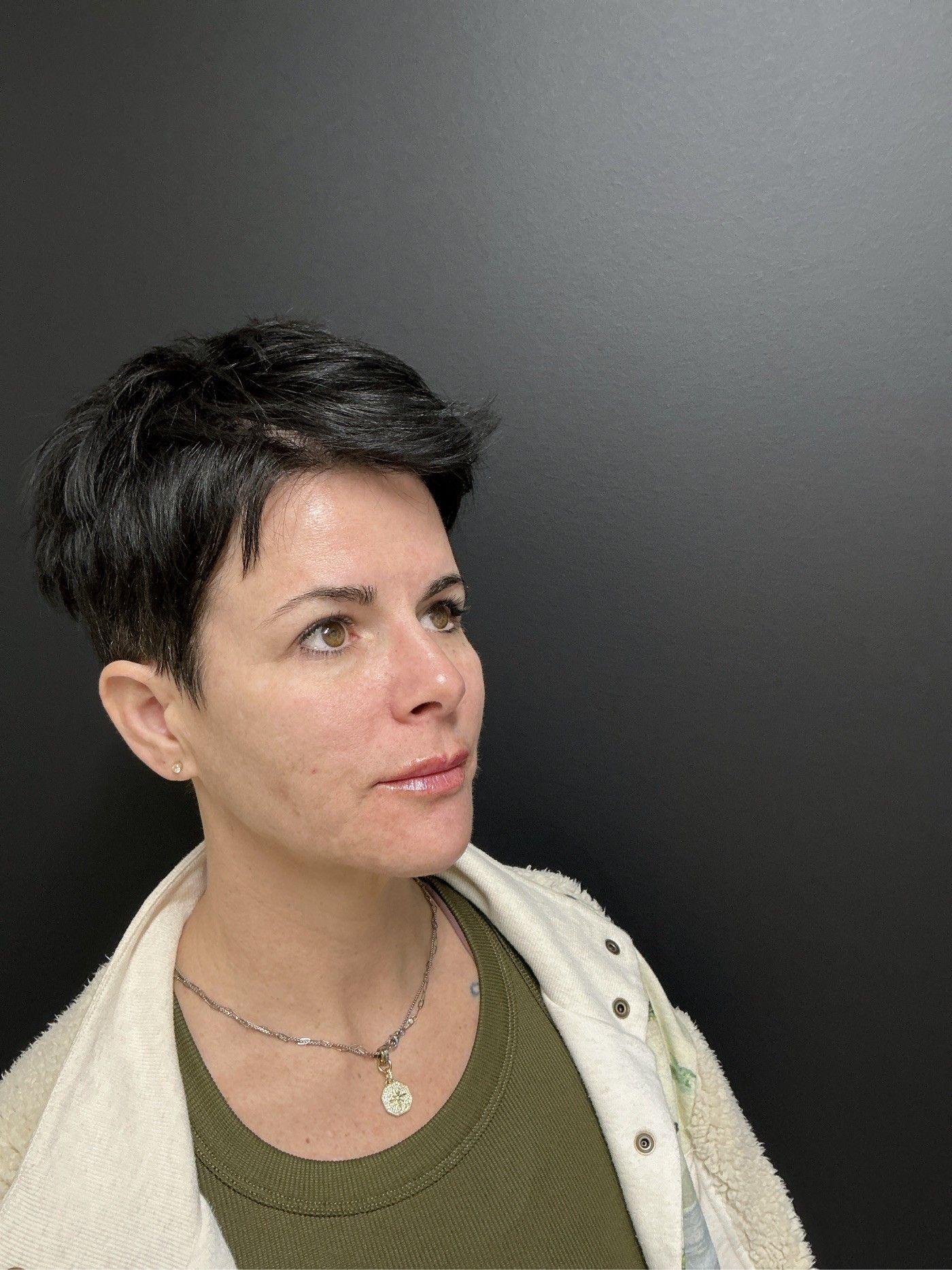 Woman with short, dark hair, wearing a green shirt and a white jacket, looking upwards, against a dark background.