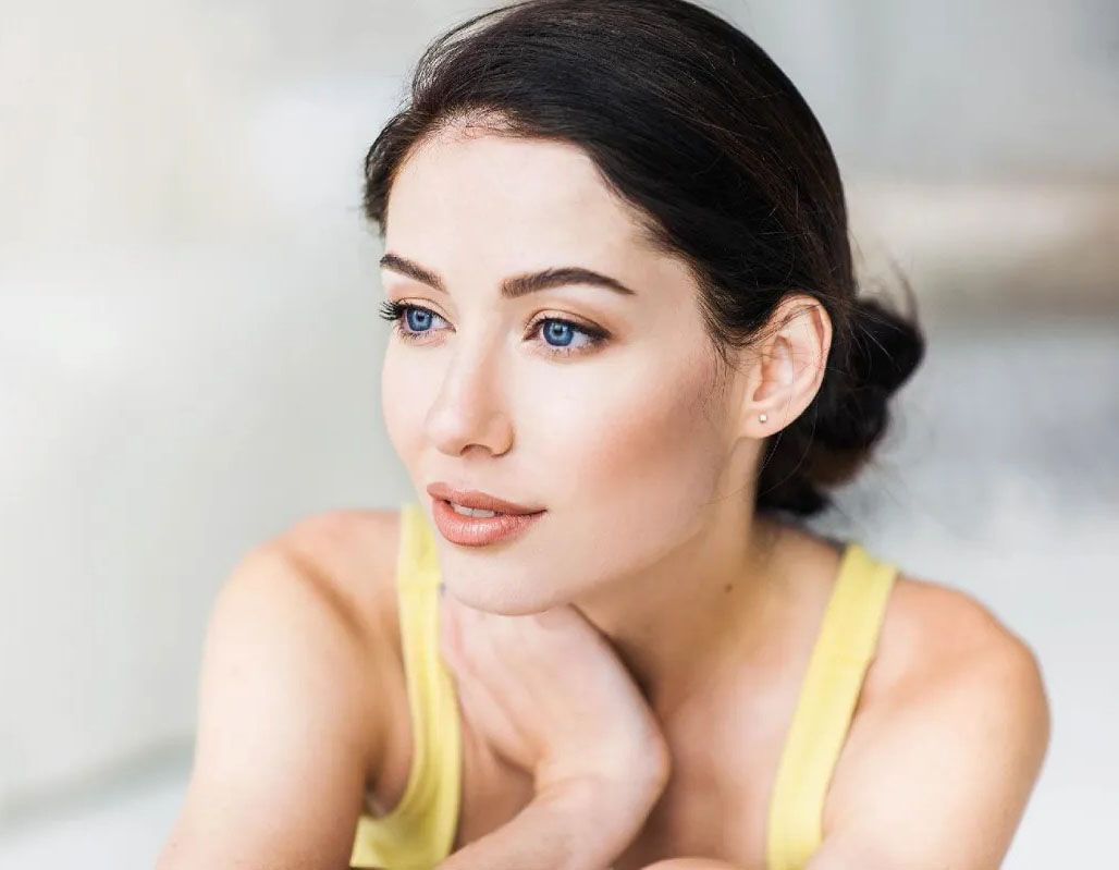 Woman with blue eyes, looking away, resting chin on hand; wearing yellow top.
