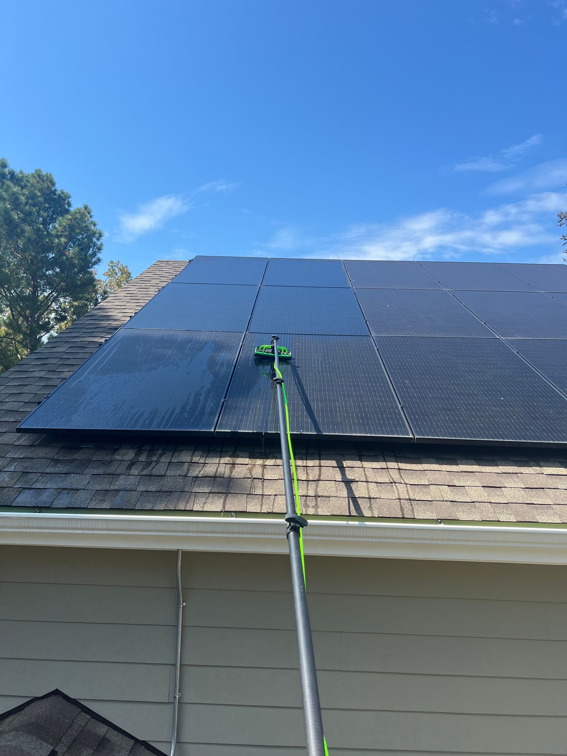 A person cleans solar panels on a roof with a long-handled brush under a bright blue sky.