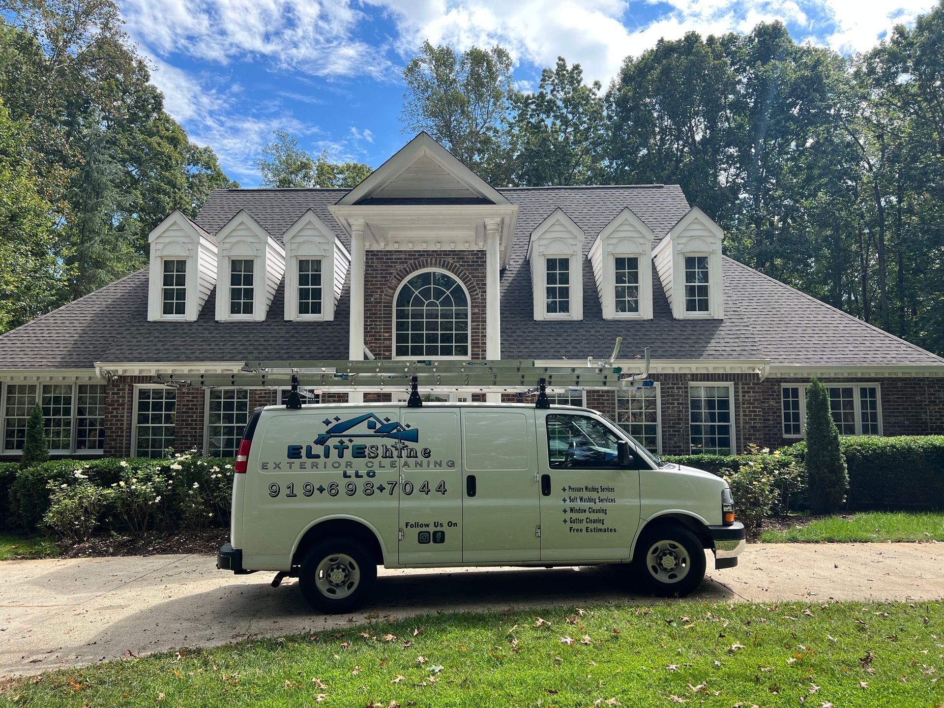 White van in front of a large brick house with dormer windows, trees in the background.