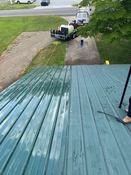 Green metal roof being pressure washed, showing cleaned vs dirty sections.