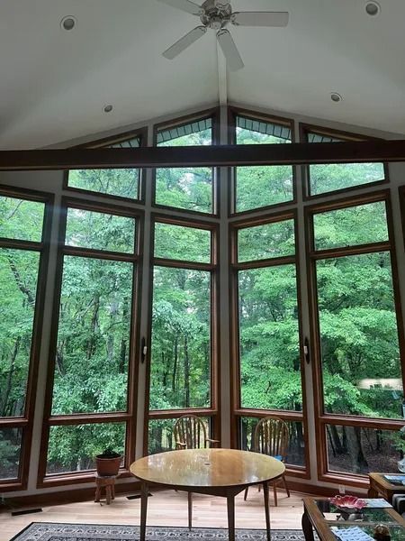Tall windows frame a lush green forest, with a wooden table and chairs inside.