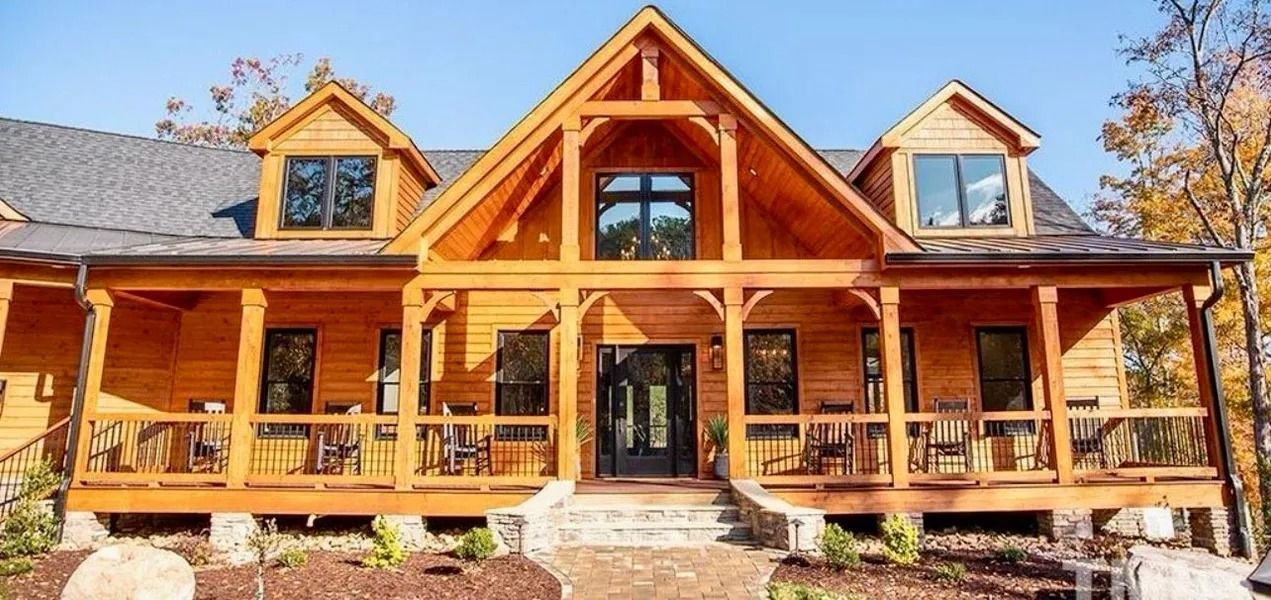 Wooden cabin with porch, dormers, and timber framing. Surrounded by landscaping, trees, and a blue sky.