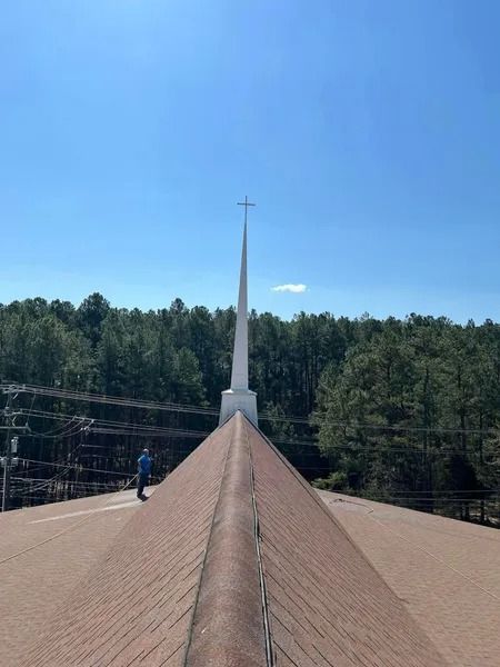 Man on church roof, pointed steeple with cross against blue sky, trees in background.