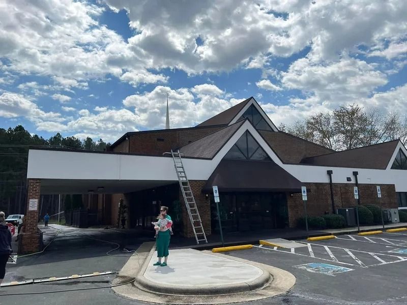 Church with a ladder on roof; a person in green stands on sidewalk under a cloudy sky.