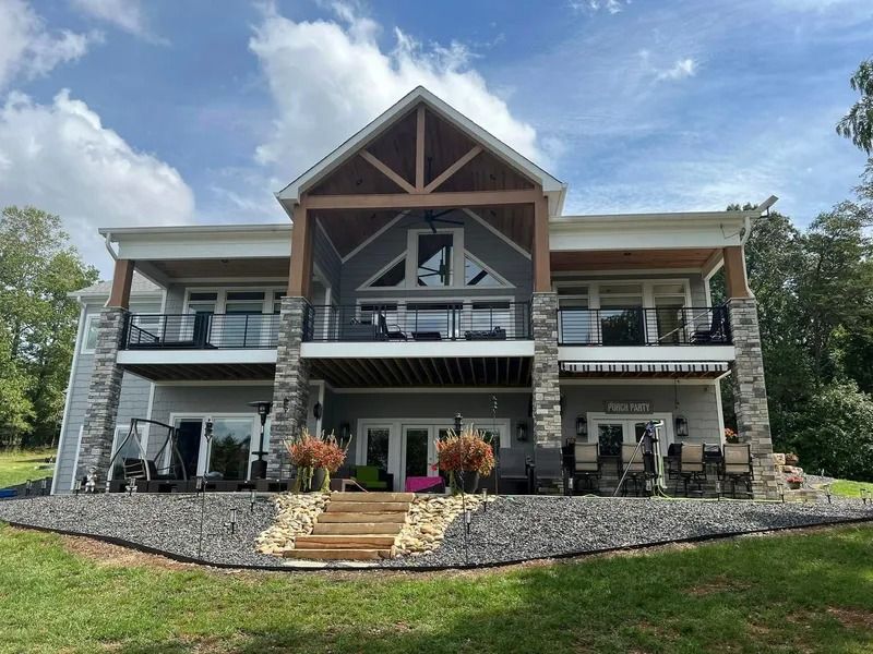 Two-story lake house with gray siding, stone accents, balconies, and a gabled entrance with wooden beams; blue sky.