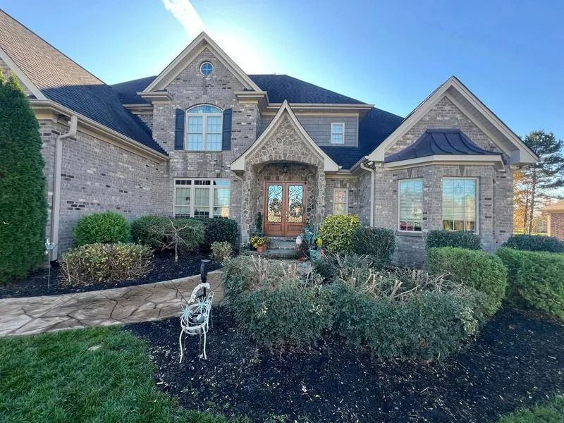 A two-story brick home with a black roof, bushes, and a stone walkway on a sunny day.