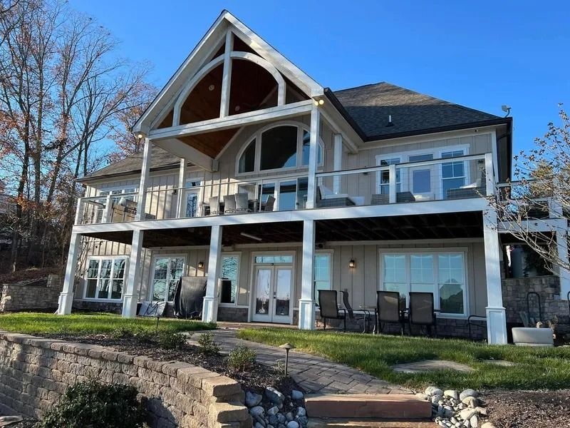 Two-story lake house with light siding, balconies, and a large arched window under a clear blue sky.