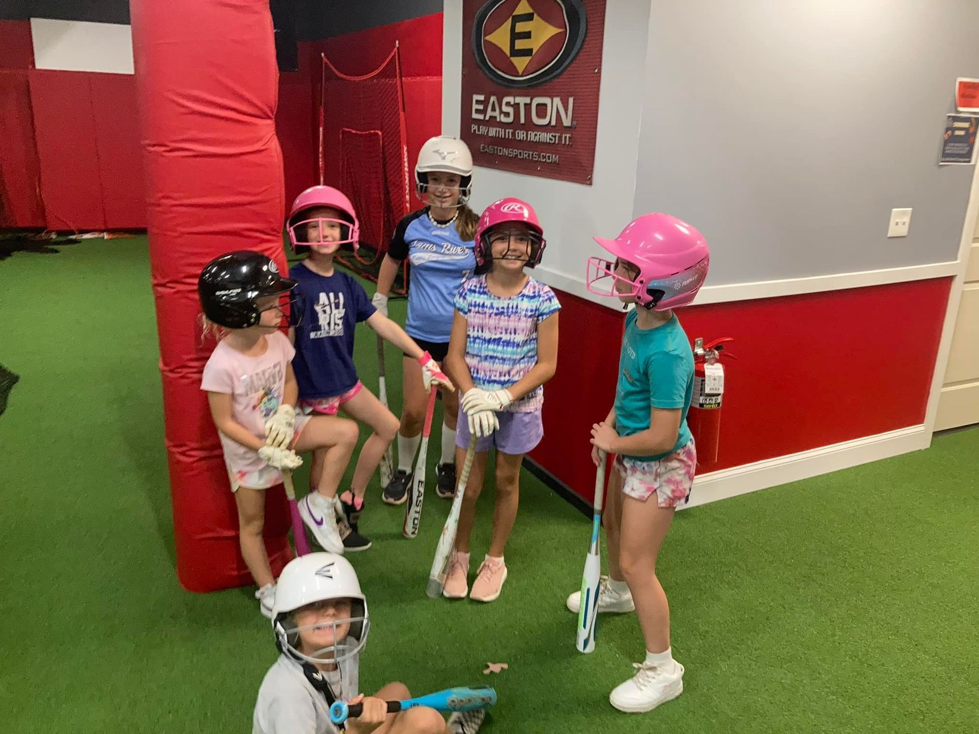Children with lacrosse sticks pose in indoor sports center wearing helmets and pads
