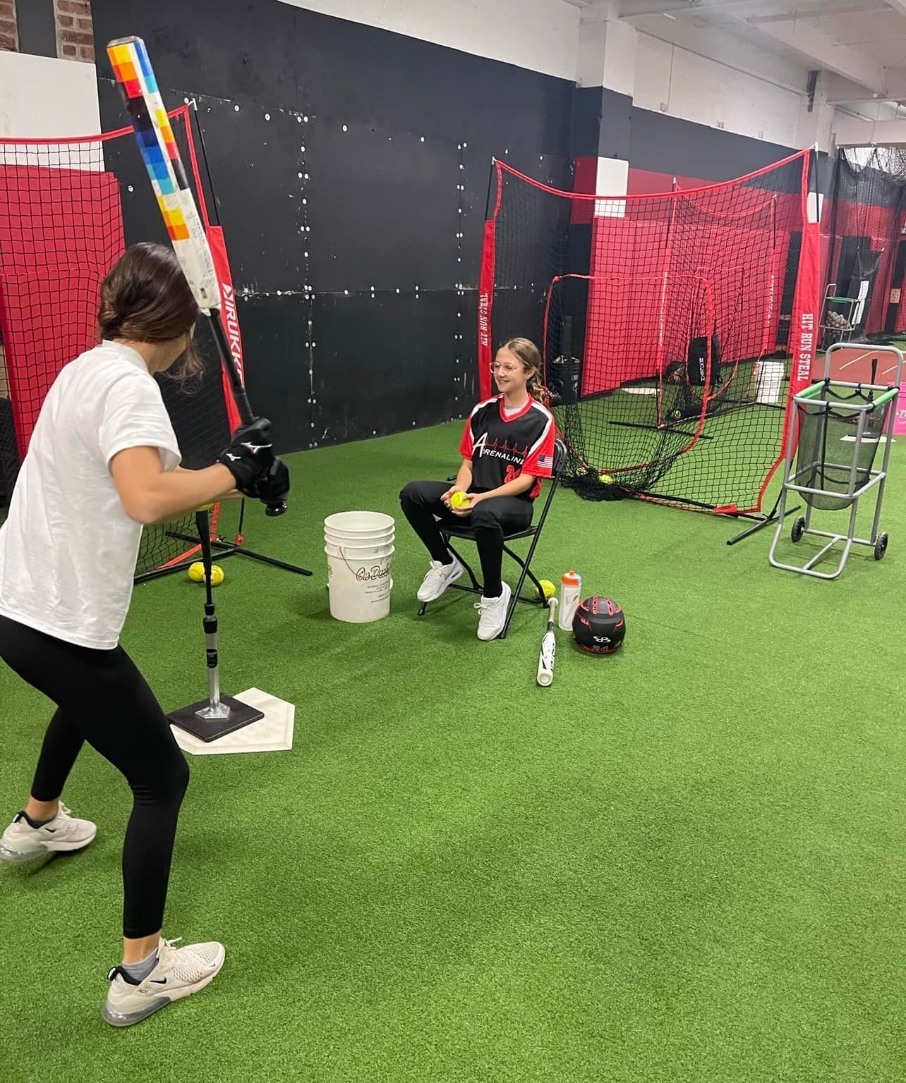 Indoor batting practice: a player swings at a tee while a coach watches in a netted training area.
