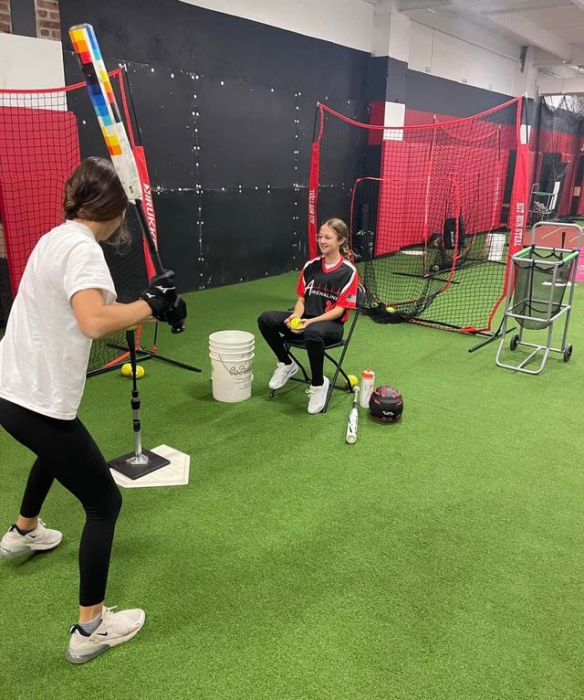 Two people in a batting cage, one pitching underhand to a seated batter with a bat and helmet.