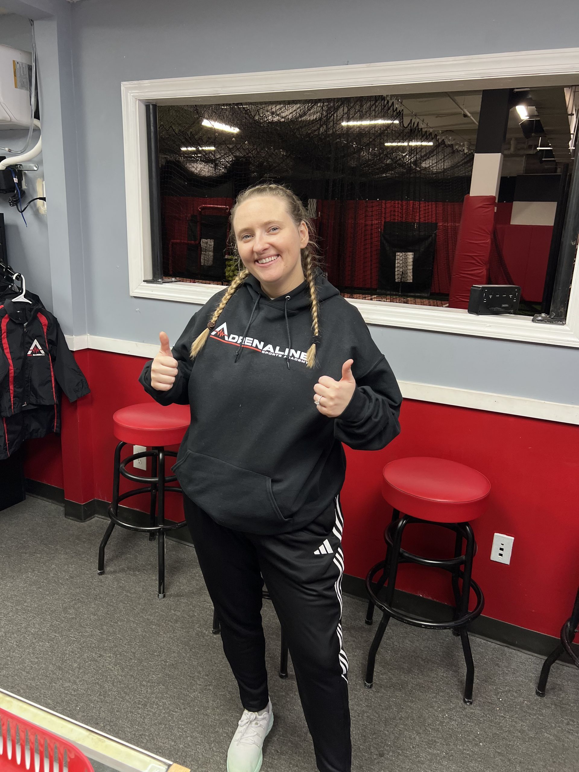 Person posing with thumbs up in a gym, wearing a black hoodie and black track pants near red stools.