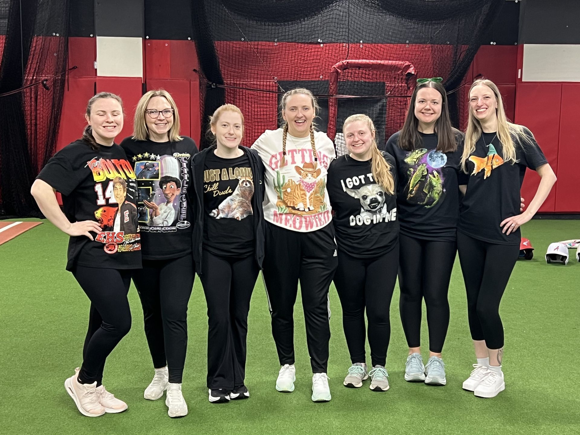 Seven people pose on indoor turf wearing black graphic T-shirts and black pants in front of netting.
