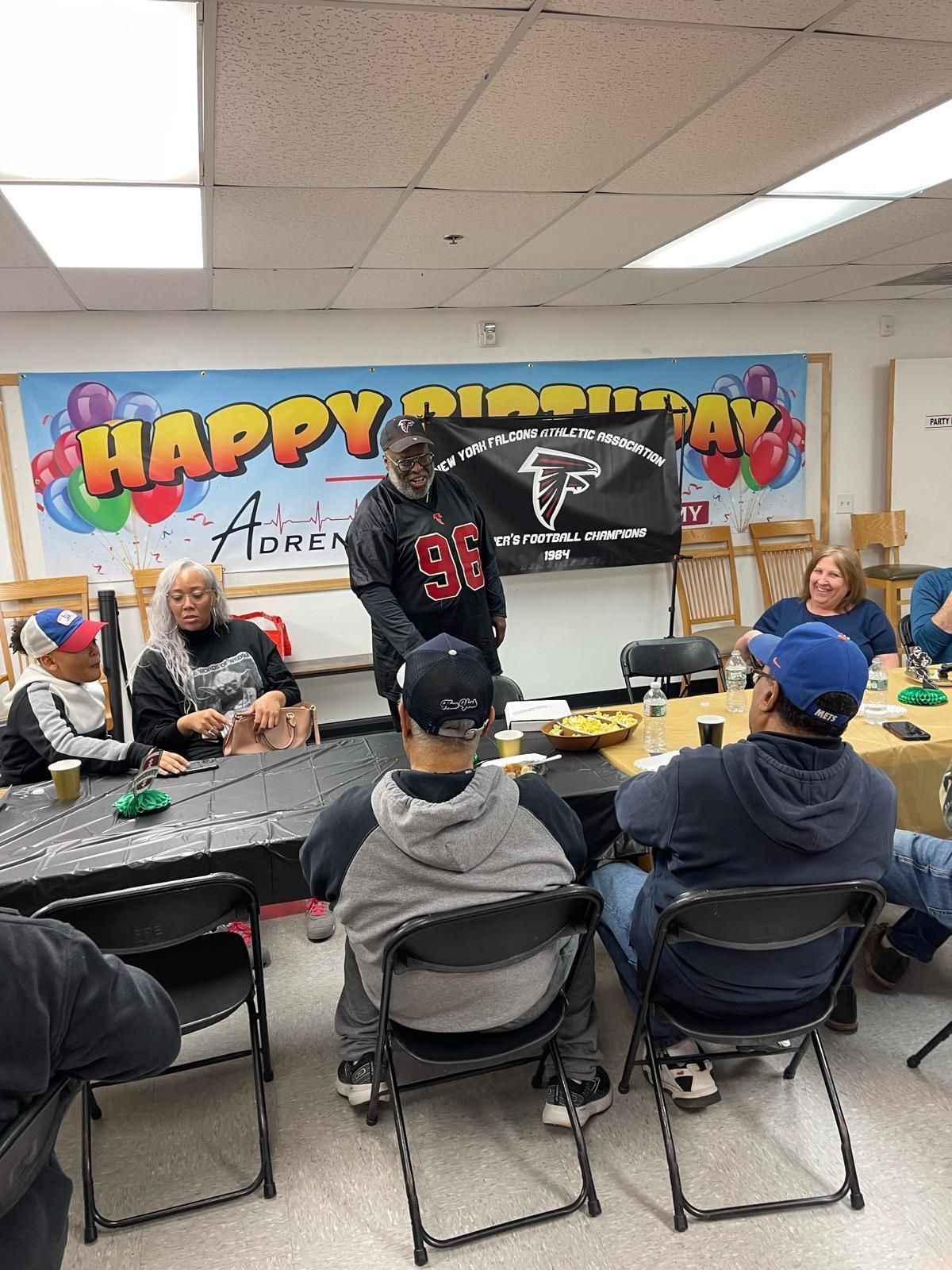 Birthday party in a decorated room, with people gathered around a table and one person presenting at the front.