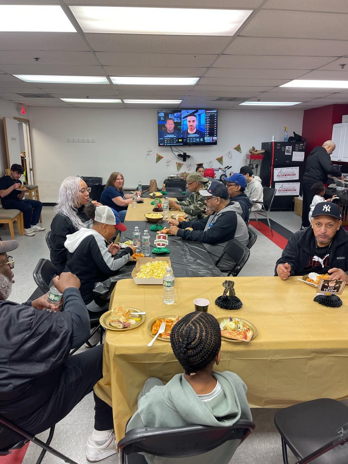 People seated around long tables in a community hall, eating together while watching TV.
